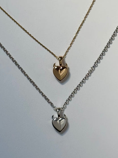 Two heart-shaped necklaces with tiny horns on top, one in gold and one in silver, displayed on a light background.