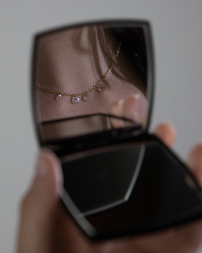 A close-up image of a diamond letter necklace with gold chain and platinum jump rings in a compact mirror, displayed on a flat surface.