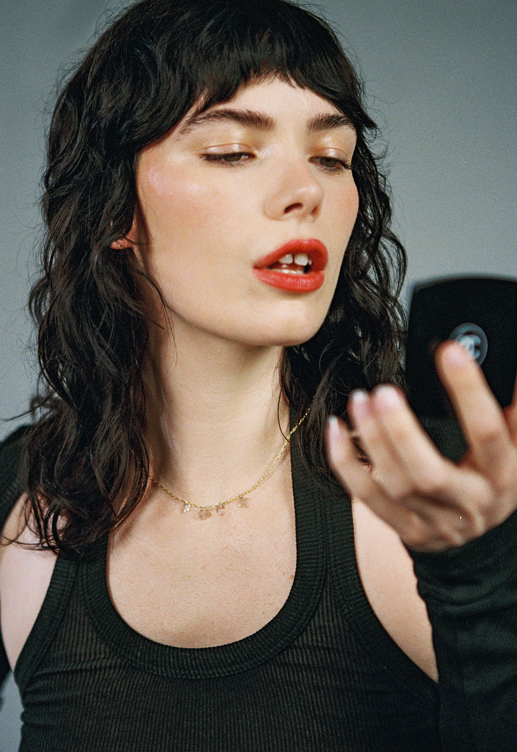 Woman with dark hair and red lipstick using a mirror wearing a diamond letter necklace against a gray background